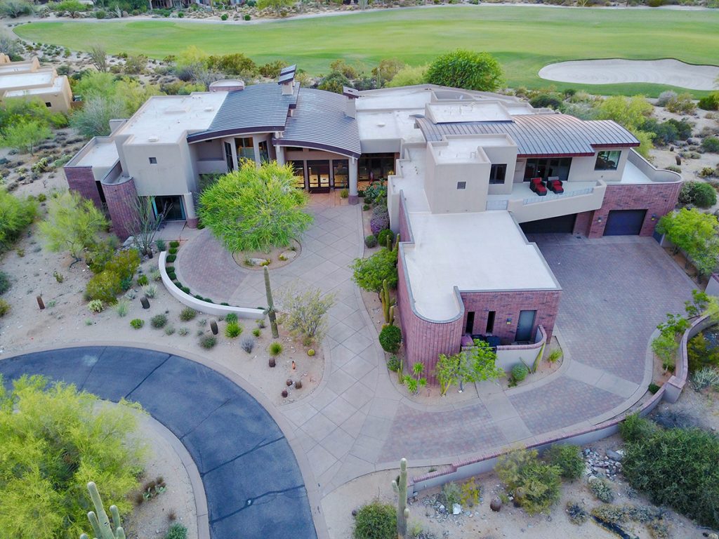 Aerial view of an opulent mansion nestled on a desert golf course, offering luxurious living with panoramic vistas of the arid landscape and lush green fairways.