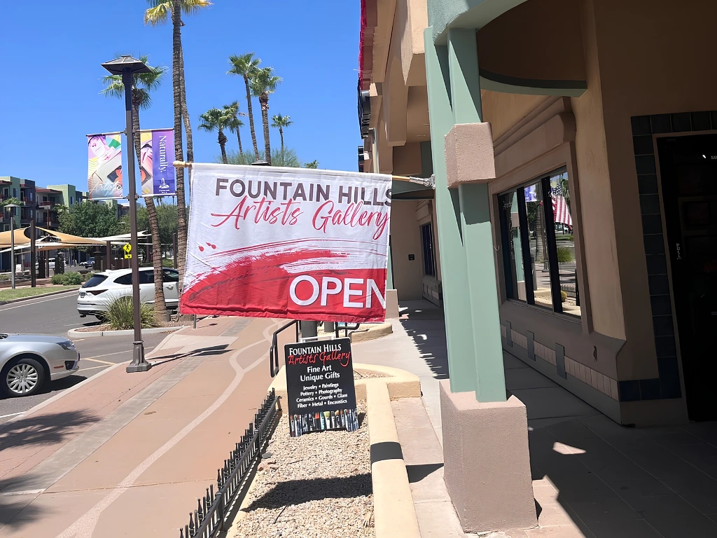 Banner for Fountain Hills Artists Gallery reading OPEN hangs in front of a storefront on a sunny palm-lined street.