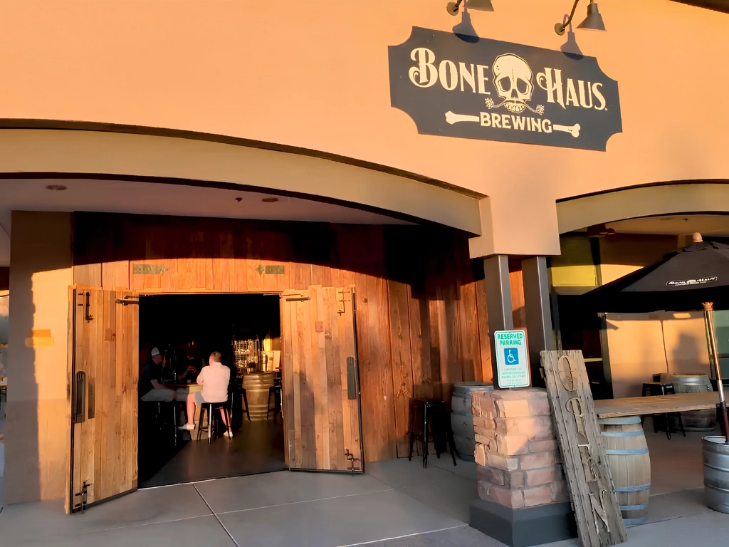 Exterior view of Bone Haus Brewing with a wooden double-door entrance and rustic barrels outside, warm sunset light.