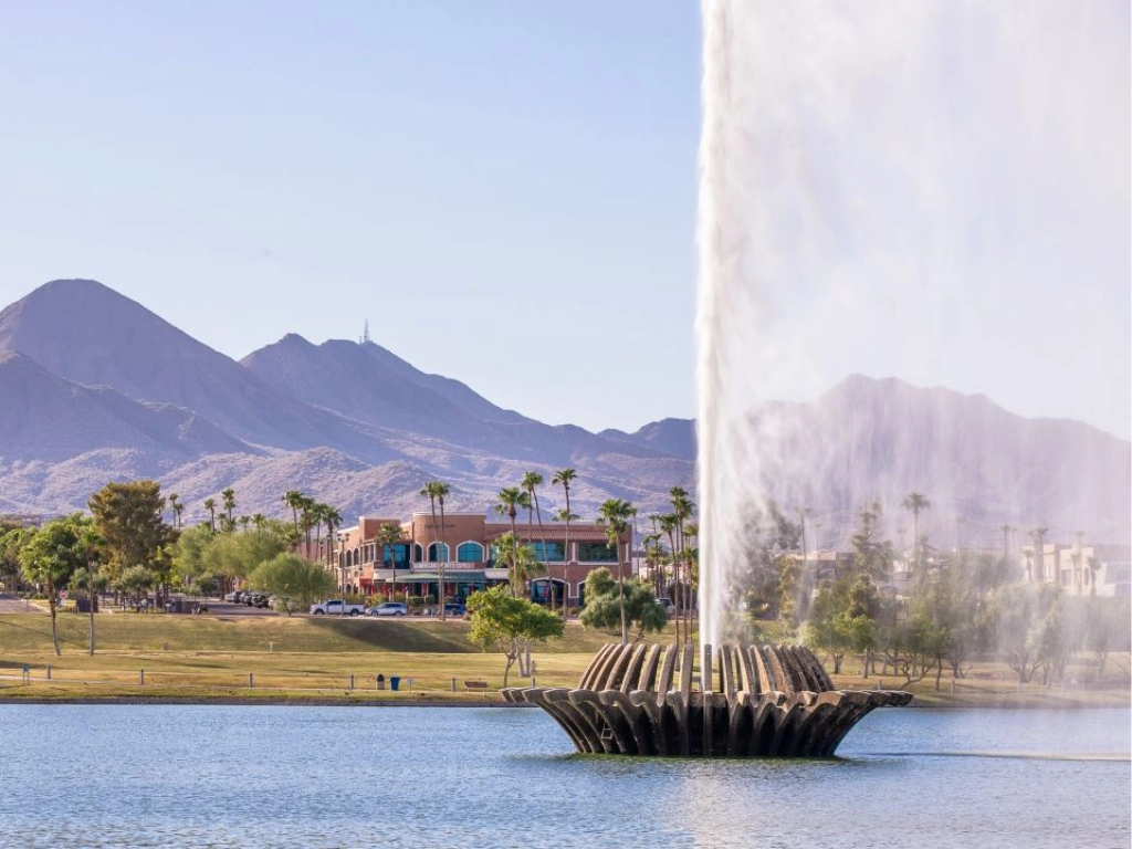 Fountain erupting from a circular basin in a lake, with palm trees, low-rise buildings, and desert mountains in the distance.