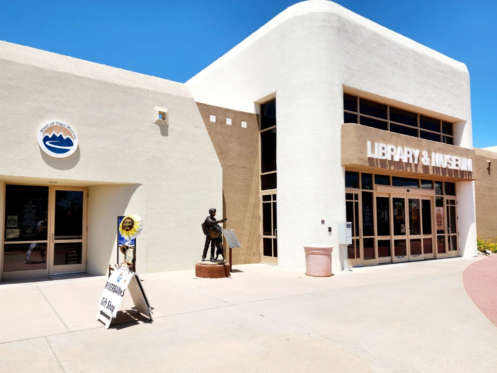 Facade of a library and museum building under a bright blue sky, with a bronze statue by the entrance and large glass doors.