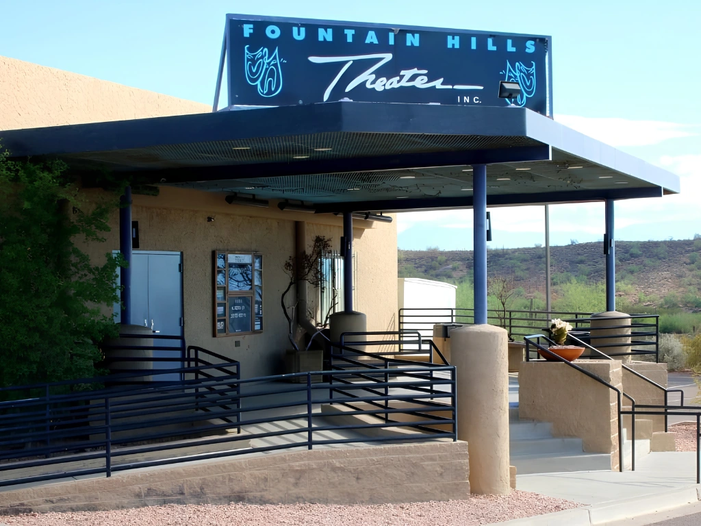 Exterior of Fountain Hills Theater with a blue sign above the entrance reading 'Fountain Hills Theater', beige stucco walls, blue railings, and desert hills in the background.